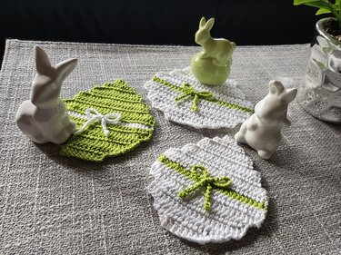 Drei gehäkelte Osterei‑Untersetzer in Grün und Weiß mit Schleifen und Hasenfiguren auf grauem Tischtuch.