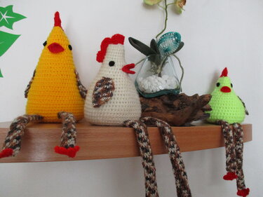 Drei gehäkelte Hühner in Gelb, Creme und Hellgrün mit roten Kämmen und langen, gemusterten Beinen auf einem Holzregal.