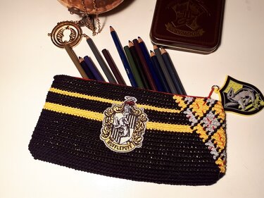 Gehäkeltes schwarzes Federmäppchen mit gelben Streifen, gesticktem wappenähnlichem Emblem und Karomuster, gefüllt mit Buntstiften.