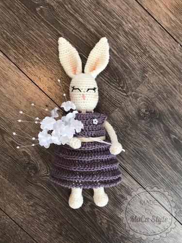 Cremefarbener gehäkelter Amigurumi-Hase in lila Kleid mit weißen Blüten auf Holzfußboden