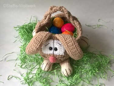 Gehäkeltes Hasenkörbchen in Hasenform, beige, gefüllt mit bunten Ostereiern und grünem Papiergras
