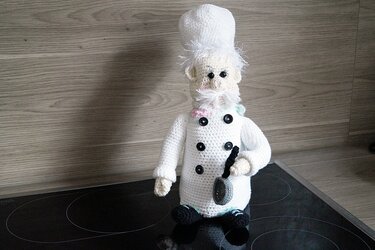 Gehäkelter Amigurumi-Koch mit weißer Kochmütze, Doppelreiher-Jacke und kleiner Pfanne.