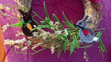 Gehäkelte braune und graue Hasen-Applikationen auf einem Strohkranz mit grünen Blättern und kleinen weißen Blüten.