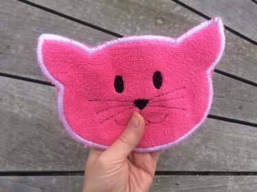 Pinkes, handgroßes Katzenmotiv aus Frottee mit gestickten Augen, Nase, Schnurrhaaren und weißer Satinkante, gehalten von einer Hand