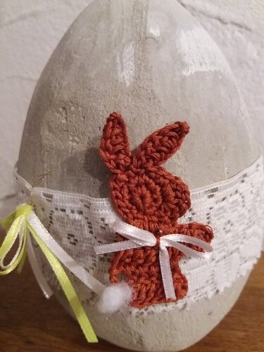 Gehäkelter rotbrauner Hase mit weißem Bommelschwanz und Schleife auf grauem Dekoei mit weißer Spitze und Bändern.