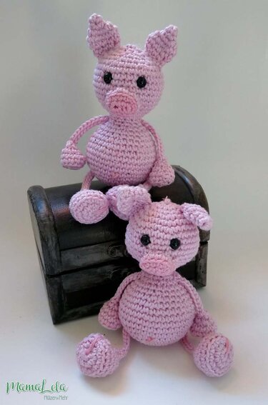 Zwei rosa gehäkelte Amigurumi-Schweinchen mit schwarzen Augen, eines auf einer dunklen Truhe sitzend