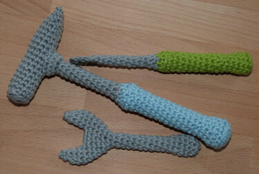 Three crocheted toy tools: grey hammer with blue handle, grey wrench, and grey screwdriver with green handle on a wood surface.