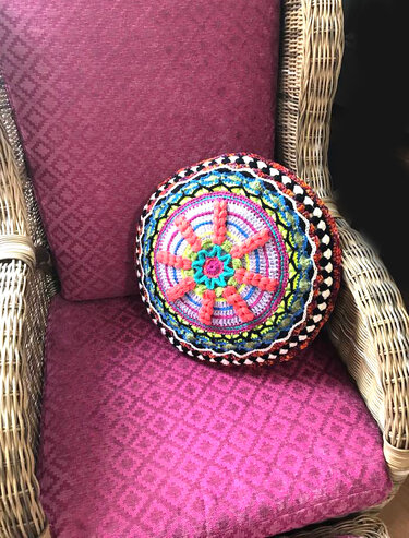 Rundes, gehäkeltes Kissen mit buntem Mandala auf einem Korbsessel mit magentafarbenem Polster