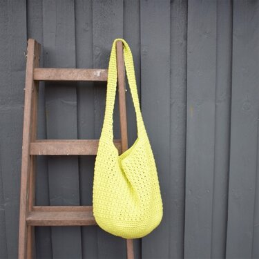 Gehäkelte gelbe Shopper-Tasche an einer Holzleiter vor dunkler Wand