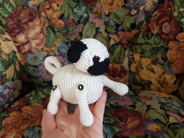 Gehäkelter weißer Amigurumi-Hund mit schwarzen Flecken, gehalten in einer Hand vor gemustertem Polster