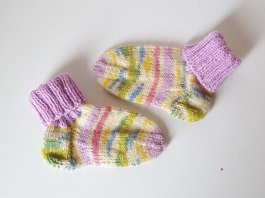 Ein Paar gestrickte Babysocken mit lila Bündchen und pastellfarbenen Streifen auf hellem Hintergrund.