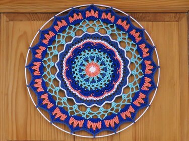 Rundes gehäkeltes Mandala in Blau-, Türkis- und Koralltönen auf weißem Ring vor Holztür