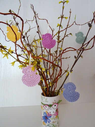 Pastellfarbene, flache gehäkelte Küken-Anhänger mit Bändern an Zweigen in einer Vase