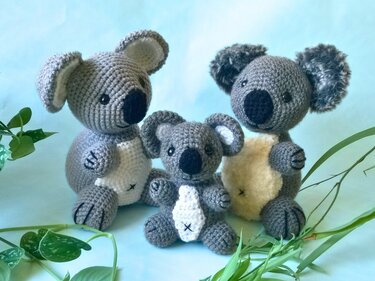 Drei gehäkelte Koalas in Grau (kleiner mittig, zwei größere seitlich) vor hellblauem Hintergrund mit grünen Blättern