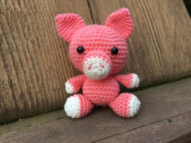 rosa gehäkeltes Amigurumi-Schwein mit weißen Pfoten und schwarzen Augen sitzt auf Holzbank
