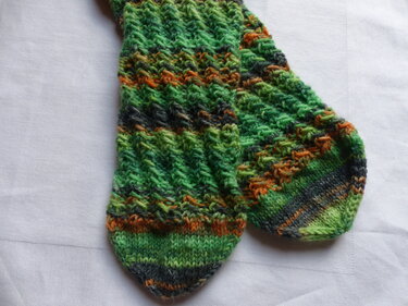 Gestricktes Paar Socken in verschiedenen Grüntönen mit orange-grauen Streifen und strukturiertem Muster, auf hellem Stoff liegend