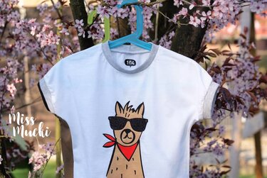 Weißes Kinder-T-Shirt mit braunem Lama-Motiv, schwarzer Sonnenbrille und rotem Halstuch vor rosa blühenden Zweigen.