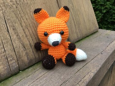 Gehäkelter Amigurumi-Fuchs in Orange, Weiß und Braun sitzt auf einer Holzbank.
