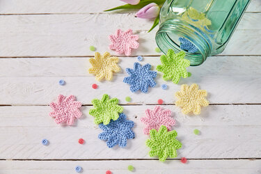 Mehrere gehäkelte kleine Blüten in Pastellfarben neben einem umgekippten Glas auf weißem Holzhintergrund, verstreute Knöpfe und eine Tulpe