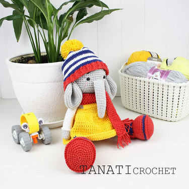 Gehäkelter Amigurumi-Elefant mit gelbem Kleid, rotem Schal und gestreifter Bommelmütze neben Garnkörben