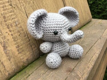 Grau gehäkelter Amigurumi-Elefant sitzend mit großen Ohren auf einer Holzbank