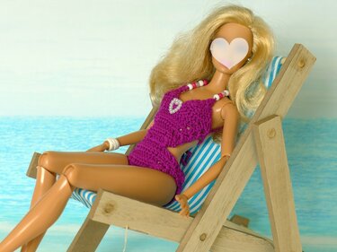 Puppe mit blonden Haaren in lila gehäkeltem Badeanzug sitzt in einem gestreiften Liegestuhl vor dem Meer.