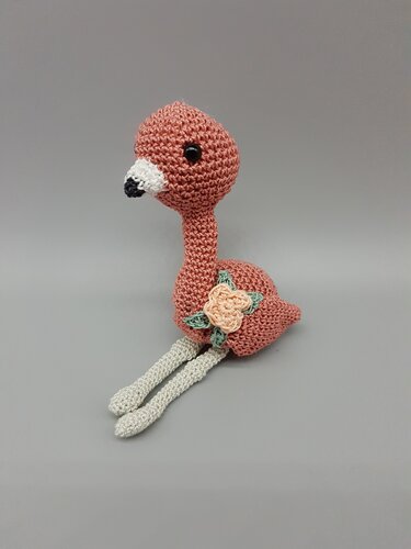 Gehäkeltes rosa Flamingo-Amigurumi mit langem Hals, hellen Beinen und Blütenapplikation am Körper