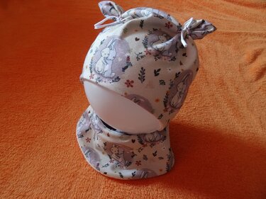 Baby-Mütze und passendes Halstuch mit Hasen- und Blumenmuster auf orangefarbenem Hintergrund.