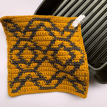 Quadratischer gehäkelter Topflappen in Senfgelb und Dunkelgrau mit grafischem Muster und weißer Aufhängeschlaufe, teilweise auf einer schwarzen Grillpfanne.