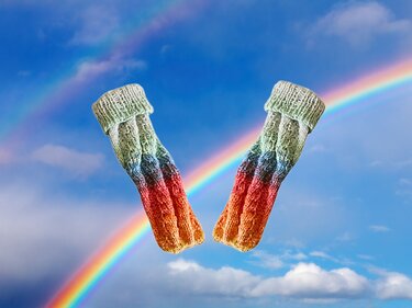 Paar gestrickter Babysocken mit Zopfmuster und Farbverlauf vor Regenbogenhimmel