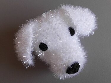 Weißer gehäkelter Hundekopf aus flauschigem Garn mit schwarzen Augen und Nase auf grauem Hintergrund.