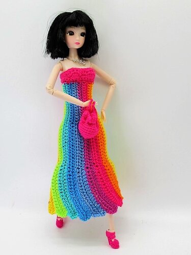 Modepuppe mit kurzem schwarzem Haar trägt ein langes, gehäkeltes Regenbogenkleid und eine kleine gehäkelte Tasche
