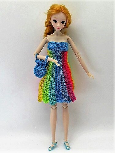 Puppe mit langem blondem Haar in gehäkeltem trägerlosem Regenbogenkleid, blauer Tasche und blauen Sandalen.