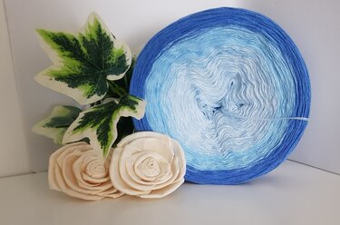 Rund gewickelter Garncake mit Farbverlauf von Dunkelblau zu Hellblau neben cremefarbenen Rosen und Efeublättern.