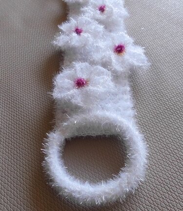 Gehäkelter weißer Handtuchhalter aus flauschiger Wimpernwolle mit fuchsiafarbenen Blüten, liegend auf beigem Untergrund