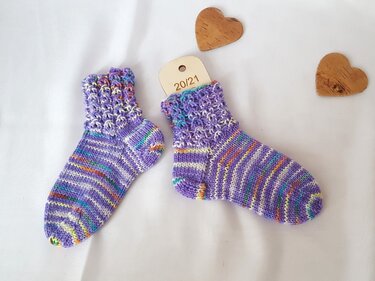 Lila, bunt gestreifte handgestrickte Babysocken mit strukturiertem Bündchen neben Holzherzen auf weißem Stoff