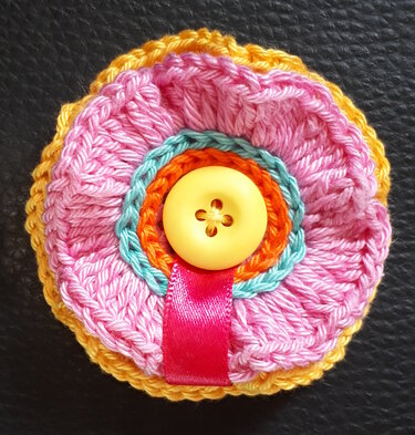 Mehrschichtige gehäkelte Blumenapplikation in Rosa, Türkis, Orange und Gelb mit gelbem Knopf und pinkem Satinband auf dunklem, strukturiertem Hintergrund.