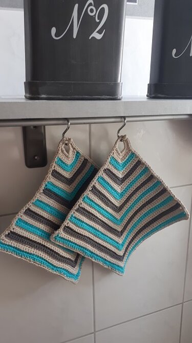 Zwei gehäkelte Topflappen mit Zickzackstreifen in Beige, Grau und Türkis hängen an Haken.