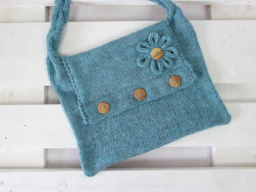 Light blue hand-knitted shoulder bag with braided strap, flower appliqué and wooden buttons.