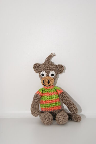 Gehäkelter brauner Amigurumi-Affe mit grün-orange gestreiftem Shirt, sitzend vor weißem Hintergrund.