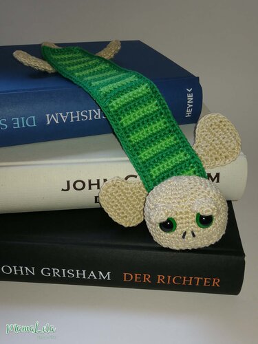 Gehäkeltes grünes Lesezeichen mit beigem Kopf und großen grünen Augen auf einem Bücherstapel.
