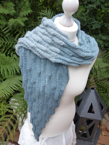 Gestricktes blaugraues Dreieckstuch mit Rippen- und Noppenmuster auf einer Schneiderbüste vor grünen Farnen.