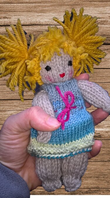 Nahaufnahme einer handgestrickten Amigurumi-Puppe mit gelben Zöpfen, blau-türkisem Kleid und pinker Schleife, gehalten in einer Hand.