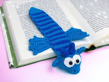 Bright blue striped crocheted dragon bookmark with large white eyes resting on an open book