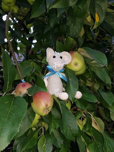 Helle, gehäkelte Amigurumi-Maus mit blau gepunkteter Schleife zwischen Blättern und Äpfeln im Apfelbaum