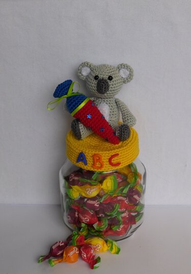 Handgehäkelter Koala mit bunter Schultüte auf gelbem gehäkeltem Deckel mit ABC-Buchstaben auf einem Glas mit Bonbons