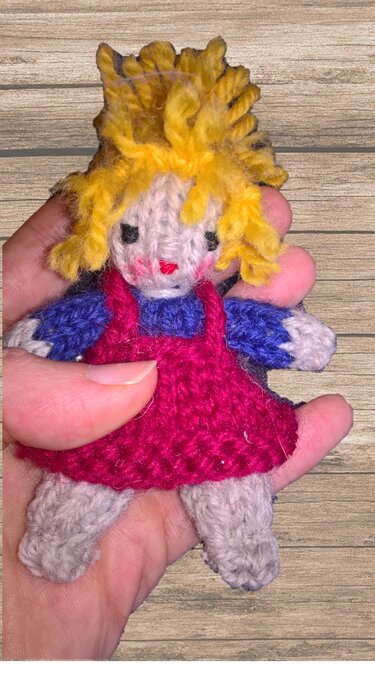 Kleine handgestrickte Amigurumi-Puppe mit pinkem Kleid, blauem Oberteil und gelbem Haar in einer Hand vor Holz.