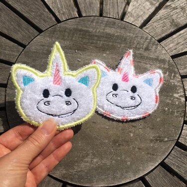 Zwei gestickte Einhorn-Aufnäher in Weiß mit gelber und rosa Umrandung, eine wird von einer Hand gehalten