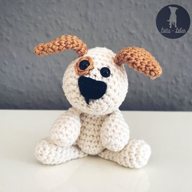 Gehäkelter sitzender Amigurumi-Hund in cremefarbenem Garn mit braunen Ohren, schwarzer Nase und braunem Augenfleck
