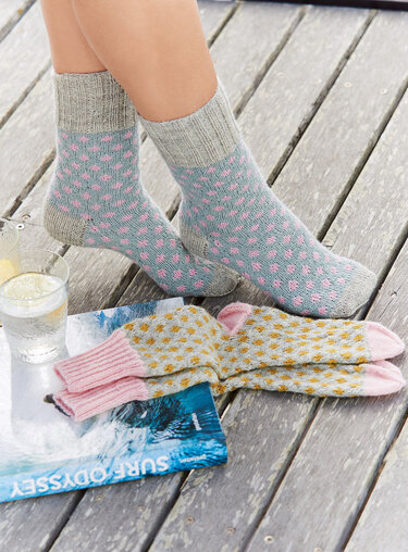 Zwei Paare gestrickter Socken auf Holzterrasse: getragenes graues Paar mit rosa Punkten, daneben ein Paar mit gelben Punkten und rosafarbenen Bündchen